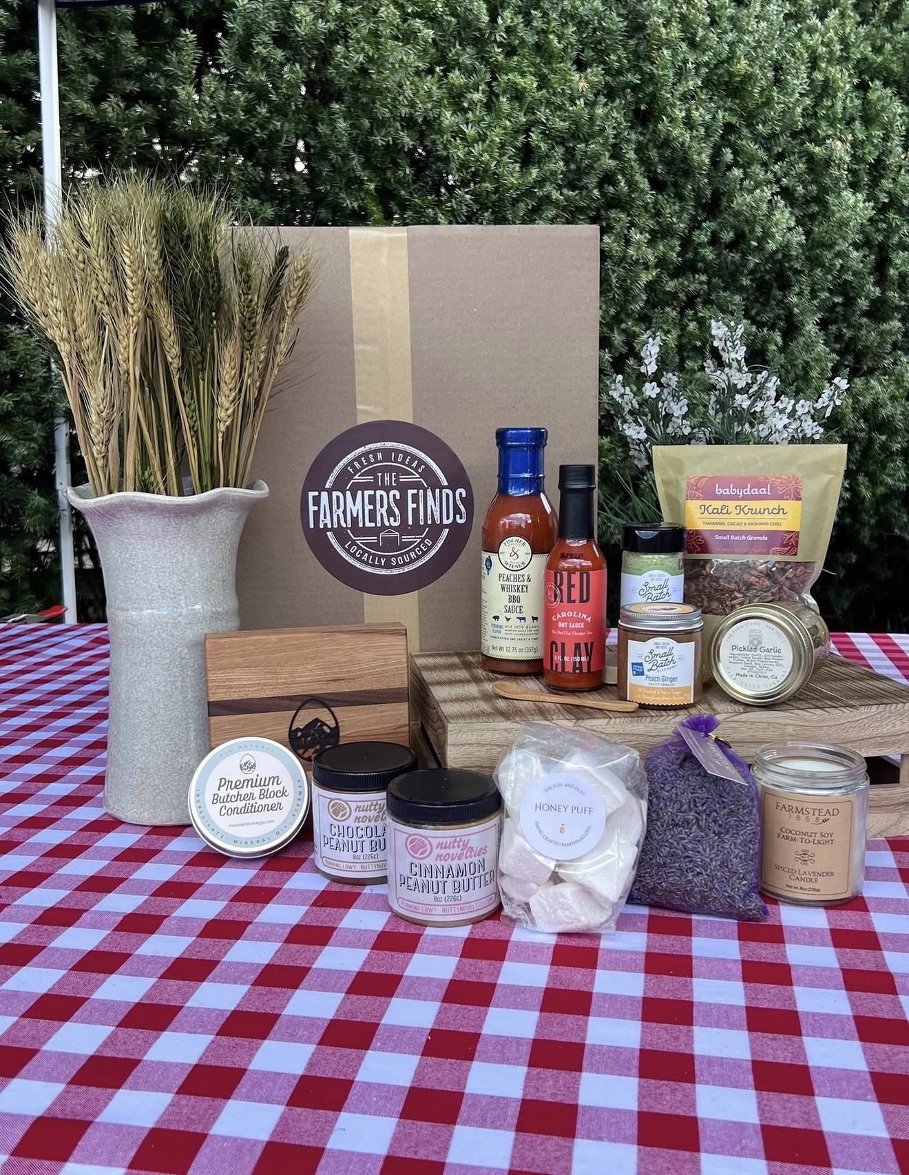 A Farmers Finds subscription box surrounded by small-batch sauces, nut butters, granola and a candle on a checkered tablecloth
